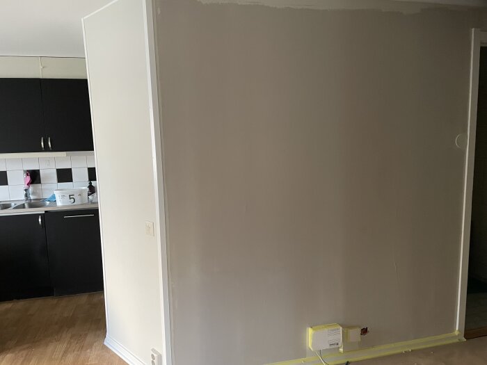 Vägg i rum med ofullständigt spacklat område mot taket, intill köksdel med svarta skåp och rutigt kakel.