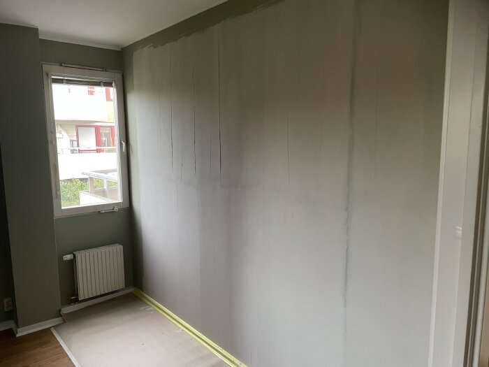 Vägg med halvklart spackelarbete där spackel inte når taket, fönster till vänster, golv täckt med skyddspapp.