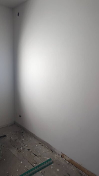 Vägg med grundmålad yta och synliga skuggbildningar från en vinkelställd arbetslampa, ojämnheter i spackling och en betonggolvsyta under.