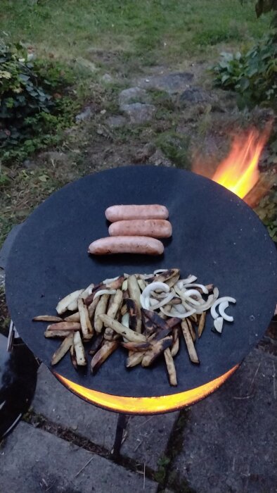Grillad fänkålssalsiccia och potatis på stekhäll över öppen eld i trädgård på kvällstid.