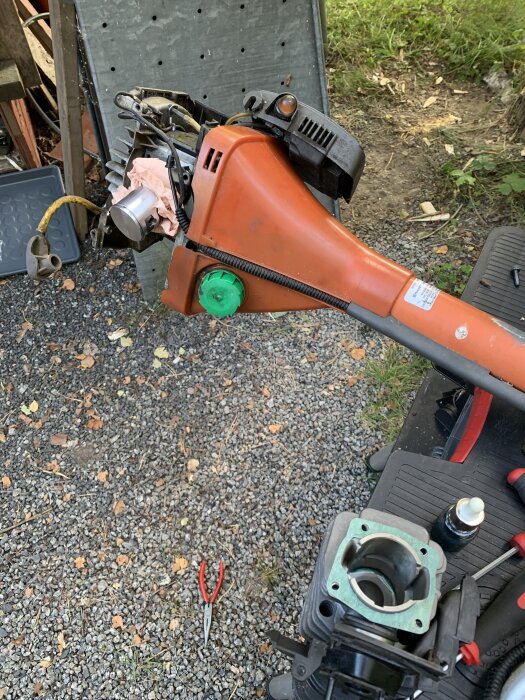 Orange röjsåg med borttagen cylinder och kolv visas, utomhus på grus. Verktyg och reservdelar ligger bredvid, inklusive röd tång och oljeflaska.