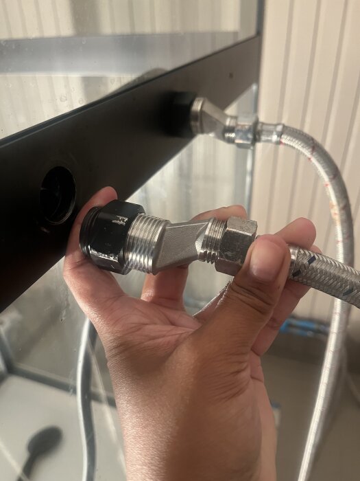 En hand håller en silvrig koppling bredvid en svart hylsa och mutter på en vägg; del av ett VVS-system i byggprojekt.