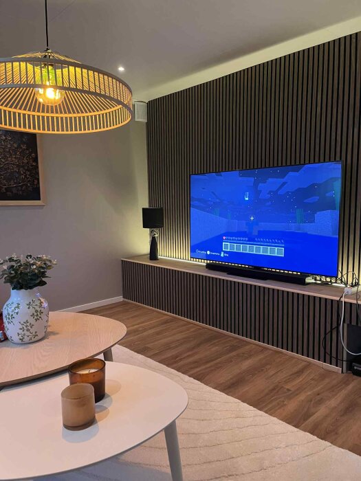 Ljust vardagsrum med en stor TV som visar ett datorspel, en stilren lampskärm, blombukett på soffbordet och en modern träpanelvägg.