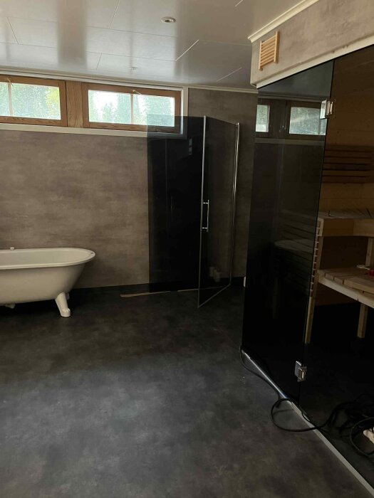 Modernt badrum under renovering med badkar, grå stenväggar och stora fönster, svart duschpanel till höger.