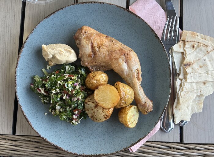 Grillad kyckling, hummus, tabbouleh, ugnsstekt potatis och libabröd på en blå tallrik.