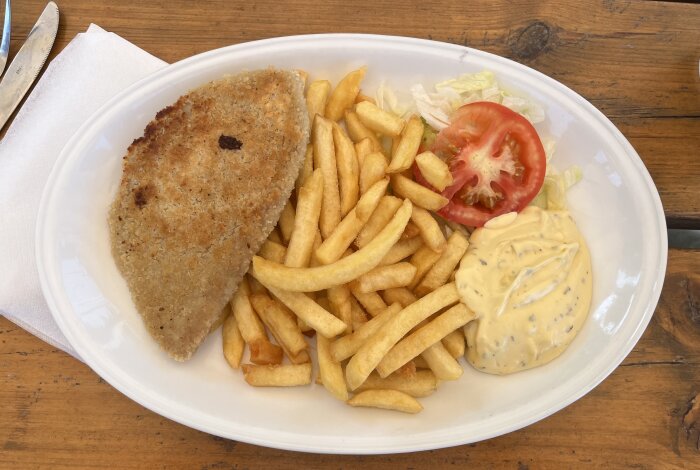 Tallrik med wienerschnitzel, pommes frites, tomatskiva, sallad och sås på ett träbord.