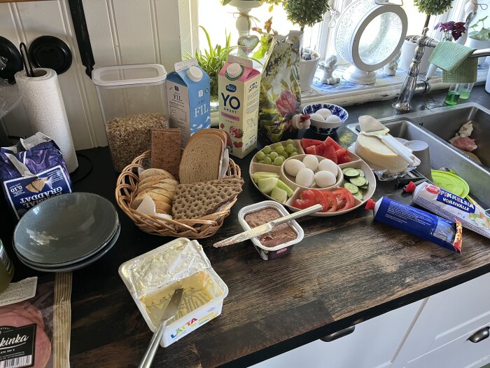 Frukostbord med bröd, fil, yoghurt, ägg, grönsaker, frukt och bredbart smör, stående i ljust kök vid diskbänk.