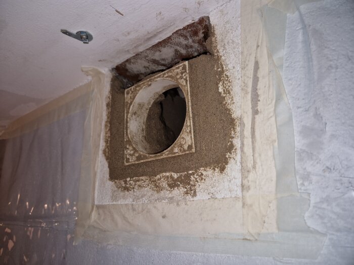 Vägg med rund ventilöppning i reparation, omgiven av bruk och tejp för försegling.