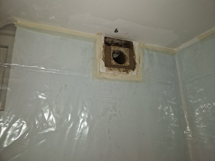 Ventiluttag i en vägg med stenskräp runt hålet, avtäckt med plast och maskeringstejp på angränsande ytor.