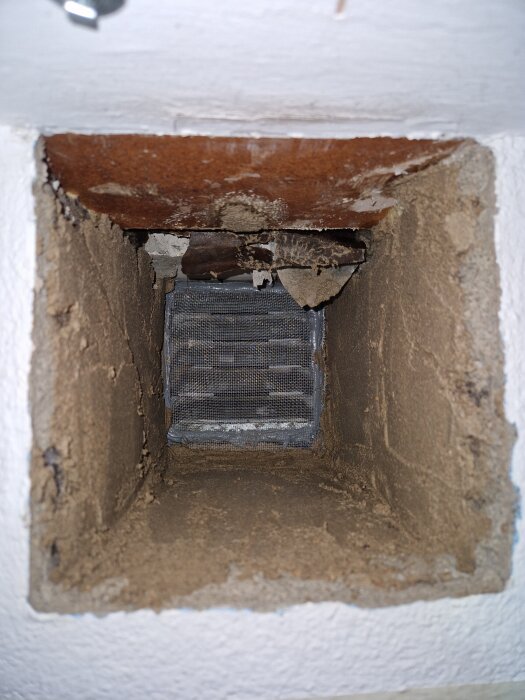Ventilationsrör med insektsnät av aluminium, omgiven av putsad mur och trä, betong inuti. Skador och justeringar synliga för renovering.
