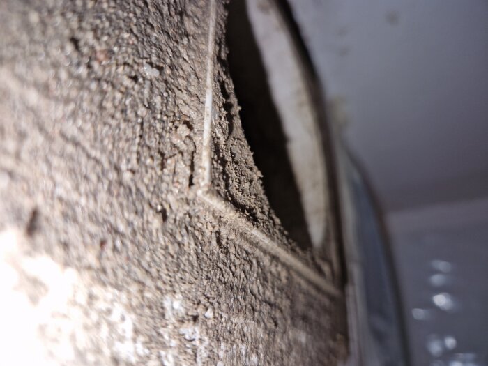 Närbild av ett grovt hål i en innervägg med ojämn yta och en cirkulär öppning, sannolikt del av ventilationsarbete.
