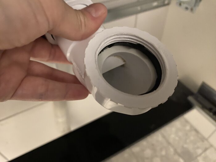 Hand håller vattenlåsets del, misstänkt packningsproblem, vit och svart komponent synlig från sidan, del av utloppsinstallation under handfat.