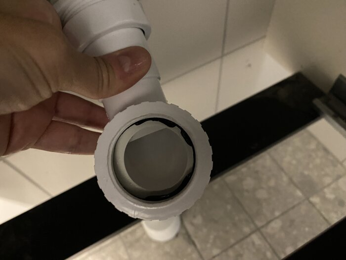 Hand håller en vit plastdel från ett vattenlås under ett handfat, med fokus på en mörk packning inuti.