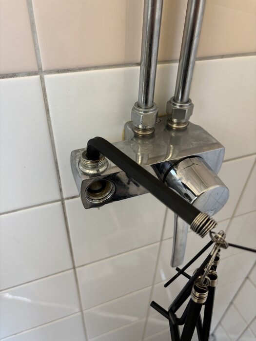 Duschblandare med synliga rör och en insexnyckel insatt i en insexmutter på övre delen av blandaren, monterade på vita kakelplattor.