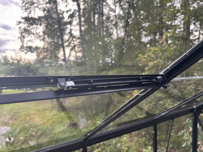 Växthusfönster med monterad öppningsarm, skruv genom skåra synlig. Bakgrund av gröna träd och gräs.