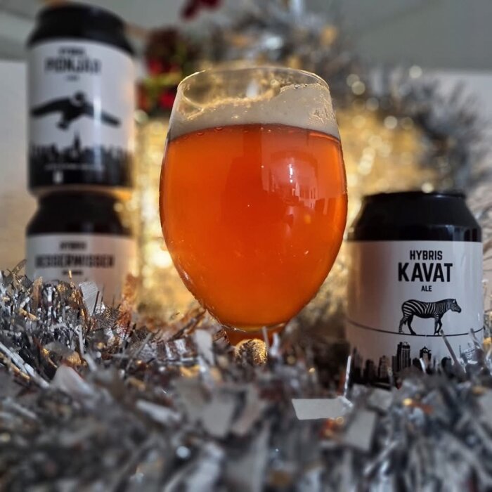 Ett glas orangefärgad öl med skumkant, omgiven av ölburkar och silverglitter i bakgrunden.