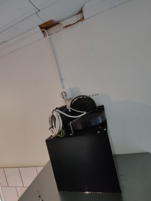 Köksfläkt sedd nerifrån, med kabel och skadad takkant ovanför, i ett kök med vitmålade väggar och tak.