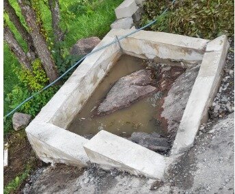 Liten rektangulär fiskdamm på berg med betongram och synlig natursten, omgiven av grönska och stenar. Vattenläckage syns i dammen.