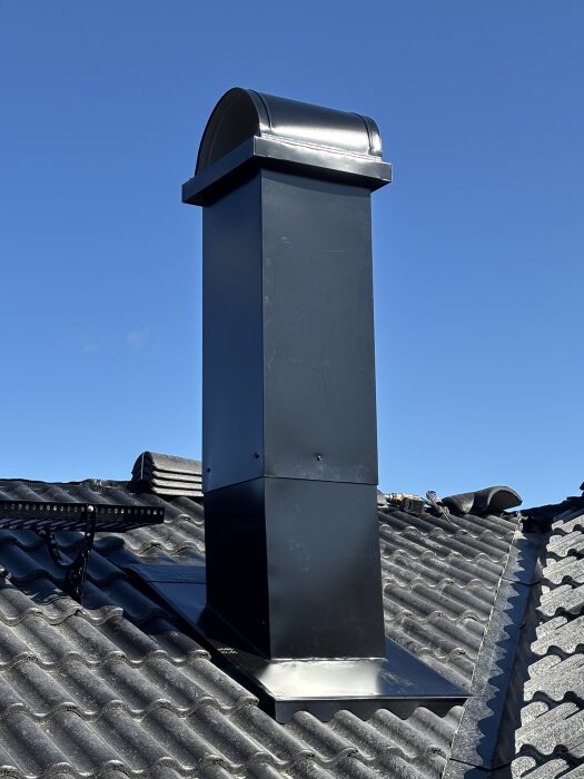 Svart plåtklädd skorsten på tak med svarta takpannor under klarblå himmel. Skorstenen är inte falsad i hörnen.