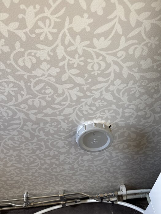Rund vit kåpa monterad på en blommig tapetvägg ovanför rör och ledningar nära golvet.