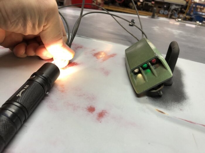Hand håller en liten lampa som lyser bredvid en grön enhet med knappar på ett bord med röda märken.