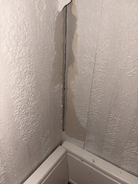 Skadad tapet i hörn på gipsvägg med synlig baksida och bortsliten yta, nära golvlist.