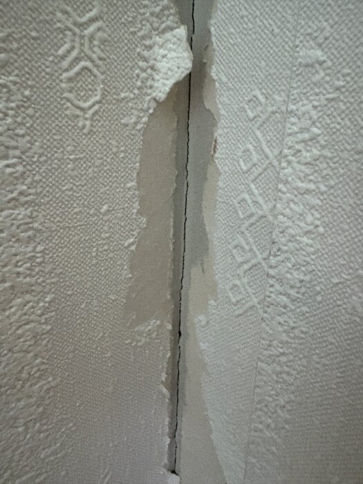 Skadad vit tapet med uppvikta kanter vid skarvar på gipsvägg; texturmönster synligt på tapeten.