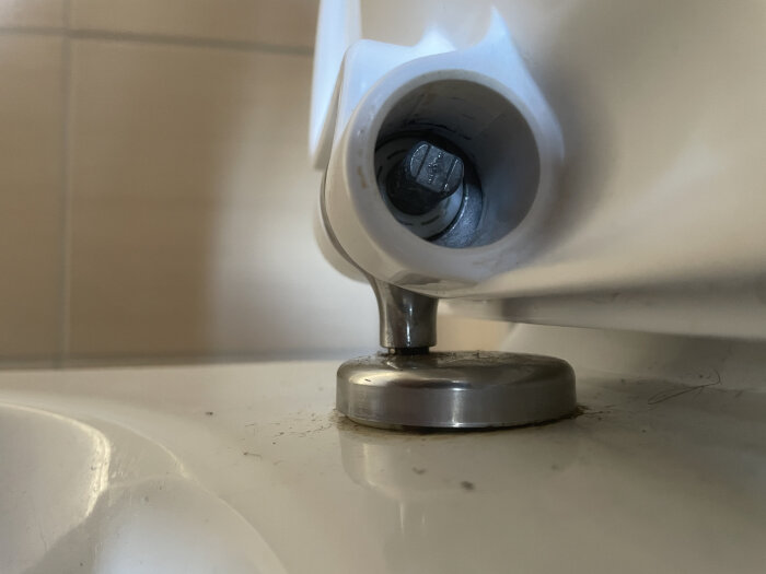 Soft-close toalettsits med saknad stoppdel och silverfärgad gångjärnsmekanism synlig på en vit yta.