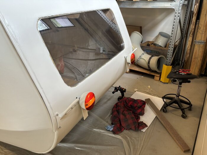Vit campingvagn under renovering med monterade baklyktor, verktyg och arbetsstol runtomkring, i ett garage.