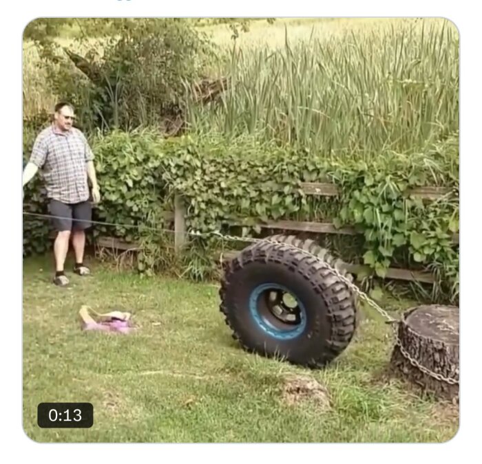 Person drar ett stort däck med en kedja fäst vid en stubbe i en trädgård med grönska i bakgrunden.
