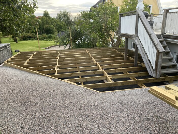 Tralldäck under konstruktion med träbjälkar, inramat av grus, bredvid en vit trappa och en gräsbevuxen trädgård i bakgrunden.