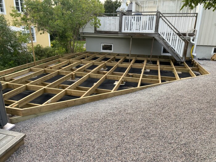 Tralldäck under byggnation med träramverk och synlig grusträdgård, samt trappa upp till veranda.