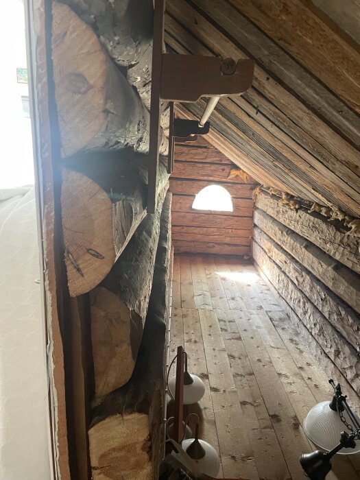 Vindsutrymme med träväggar av stockar, smala halva stockar syns i väggen. Rundat fönster och trägolv. Lampor och möbler delvis synliga.