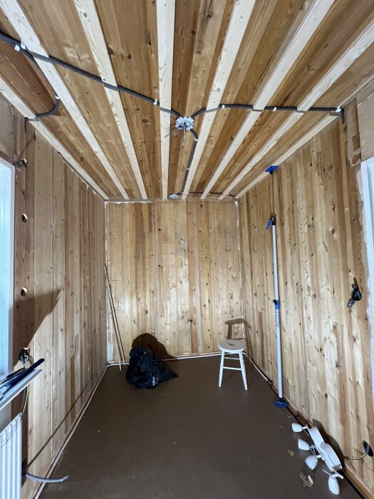Ett rum under renovering med trätak och träväggar, en flexslang längs taket, en stol, svarta säckar och verktyg på golvet.