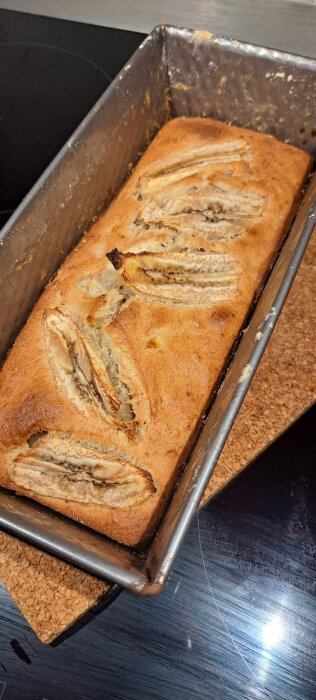 Bananbröd i metallform på en korkunderlägg, nybakat med skivad banan på toppen.