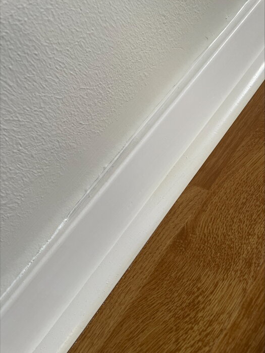 Vägg med vita tapeter och trälist i närbild, synliga luftbubblor och ojämnheter vid tapetskarven ovanför listen.