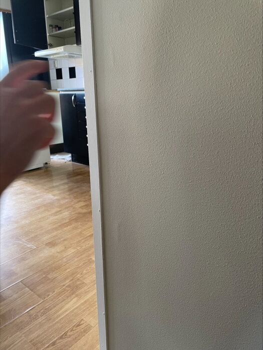 Närbild på en vägg vid en dörrlist med synliga vertikala linjer och antydan till luftbubblor under tapeten i ett kök. En hand syns i vänster kant.