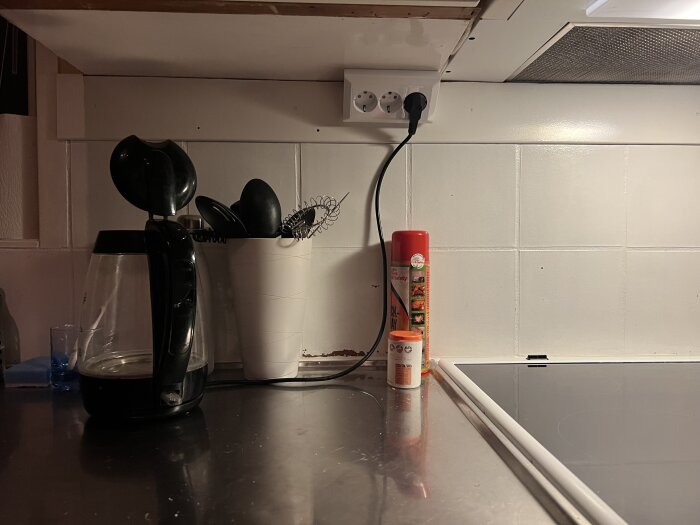 Köksbänk med vattenkokare, behållare med köksredskap, sprejburk och nytt 3-vägsuttag ovanför spishällen.