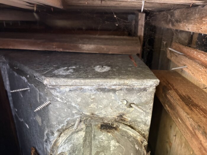 Skruvhål i plåtränna under ett tak, med synliga skruvar och vattendroppe vid ett hål, omgivna av träbjälkar. Möjligen problem med takavrinning.