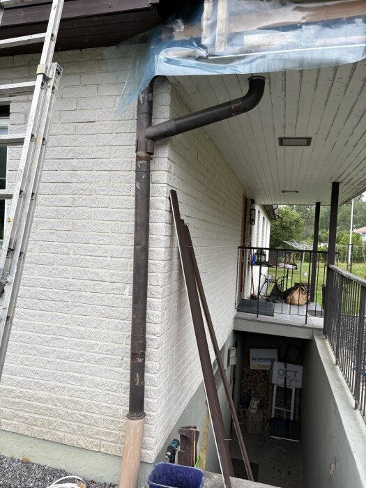 Hörn av hus med stuprör, stege lutad mot vit tegelvägg, farstukvist med tak och plastränndal synliga.