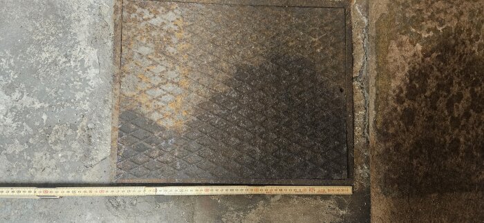 Rostigt brunnslock i ett garage, fyrkantigt och mönstrat, med ett måttband intill för storleksreferens.