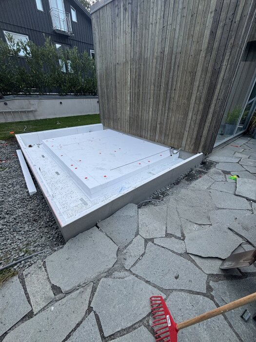 Cellplastisolerad grund bredvid träpanel på ett hus, omgiven av stengång och grus, med en kratta i förgrunden.