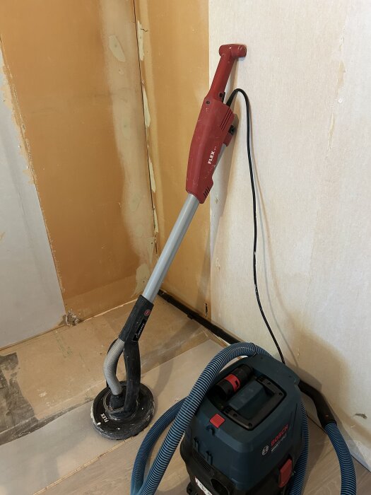 Verktyget Flex GE5 står lutat mot en vägg i ett renoveringsområde, kopplat till en Bosch dammsugare.