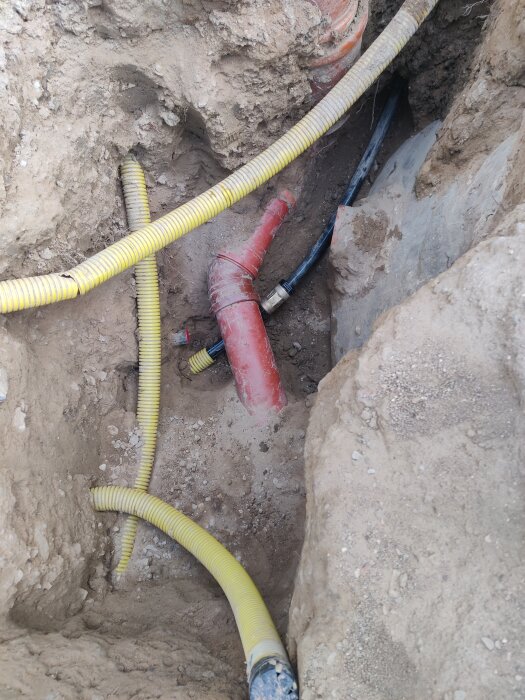 Grävd grop med röda och gula rör i jordig omgivning, visar pågående arbete med vattenrörsinstallation under mark.