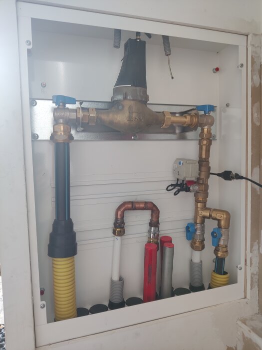Vattenmätare och rörsystem i ett skåp, med olika kopplingar och ventiler synliga. Installation för omkoppling av inkommande vattenledning.