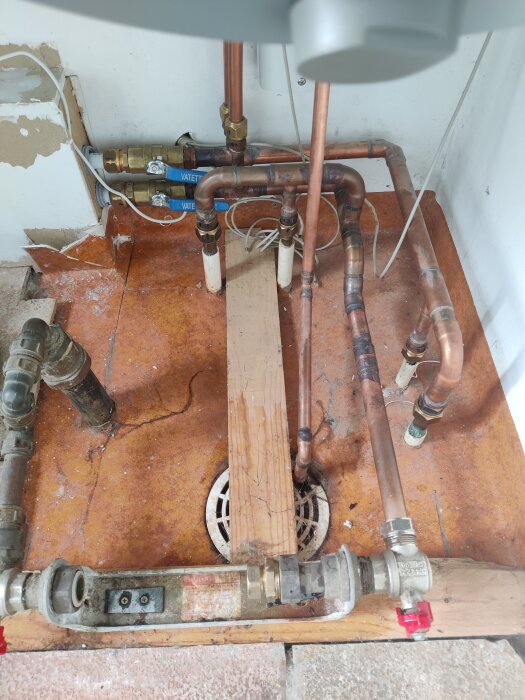 Rörsystem för vatteninstallation i en grop; nya kopplingar och vattenmätare är monterade. Ventiler med blå etiketter och rör av koppar syns tydligt.