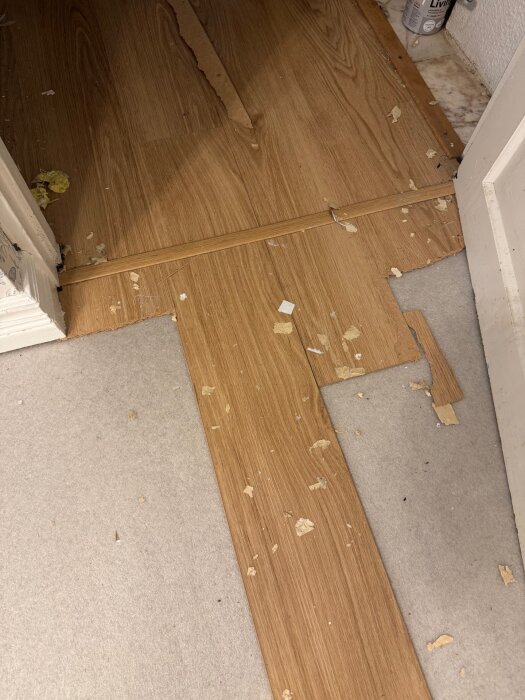 Delvis borttaget trälaminatgolv vid en dörröppning med synliga skarvar och skräp på betonggolvet.