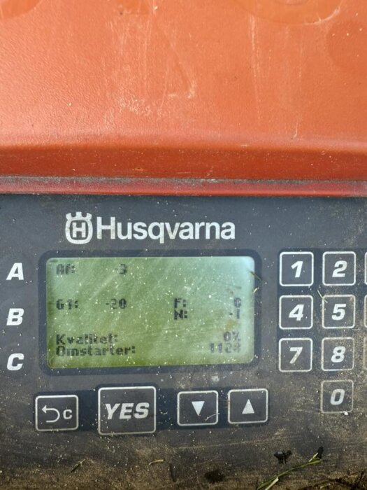 Skärm på Husqvarna Automower visar signalkvalitet 0% och omstart 1128 gånger, med en knappsats och smuts på omgivande yta.