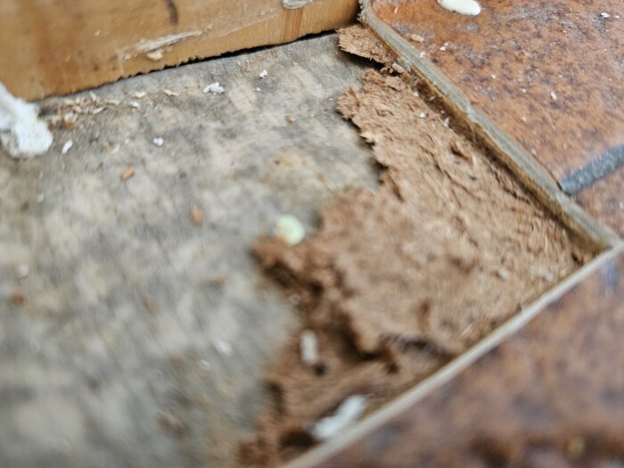 Närbild av gammalt golv med plastmatta, träfiberskiva och filtaktigt lager över trägolv. Finns risk för asbest i ett trähus från 60-talet.