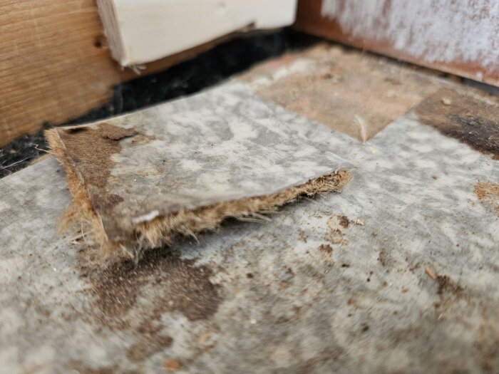 Två gamla plastmattor ovanpå varandra, den övre på en träfiberskiva och den undre på ett luddigt material, i ett trähus från 60-talet.